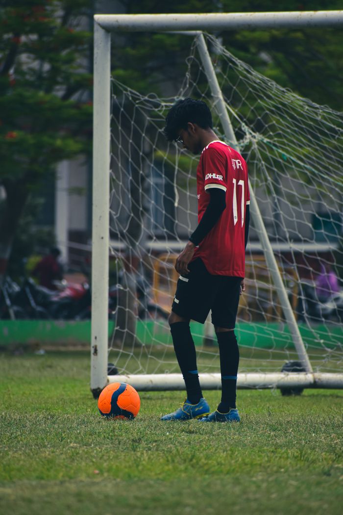 Teen soccer player in red jersey near goalpost on a sunny day in India.
